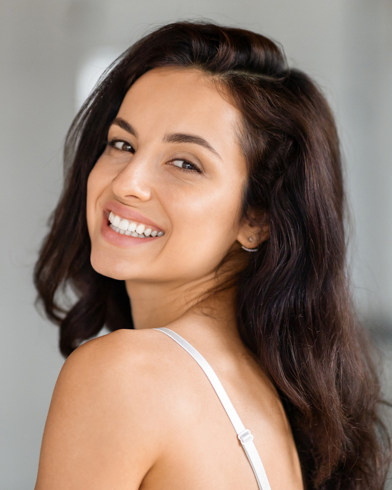 Smiling woman looking over her shoulder, long dark hair, bright teeth, natural portrait with white camisole strap