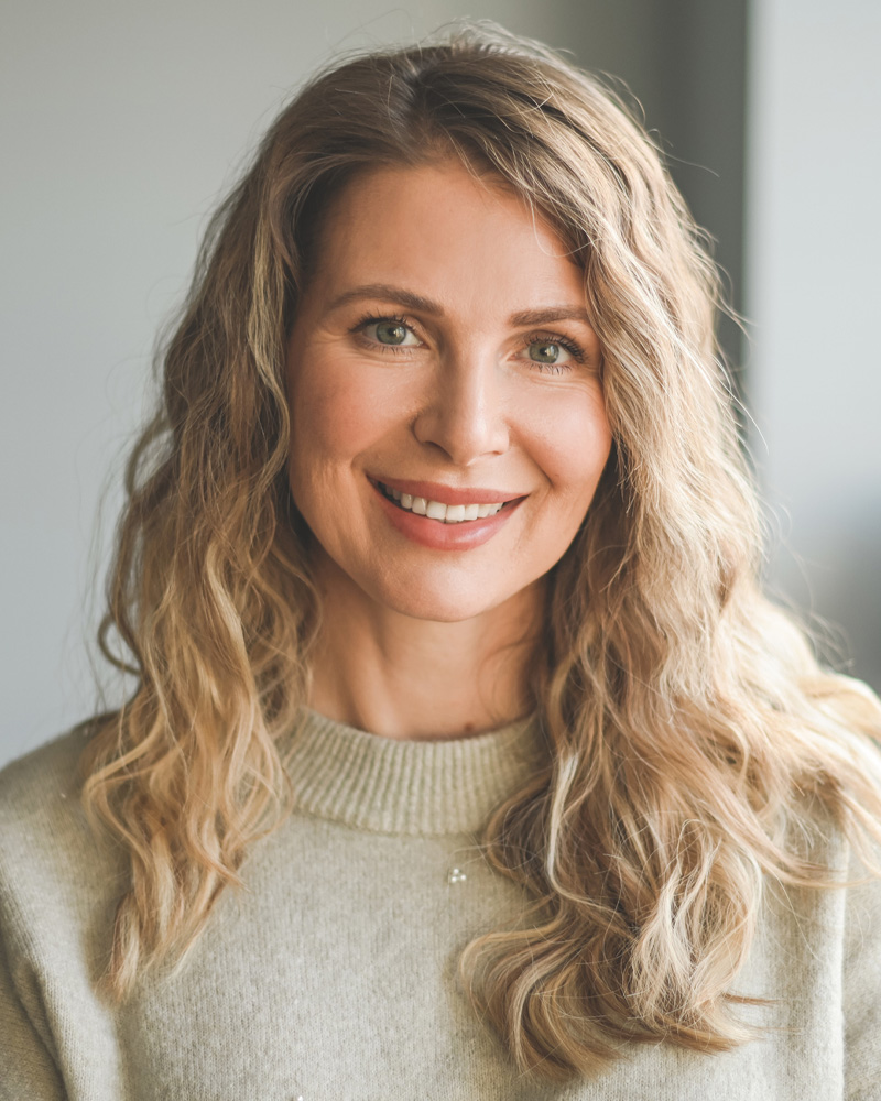 Smiling woman portrait with wavy blonde hair wearing a beige sweater, natural light headshot, warm friendly expression