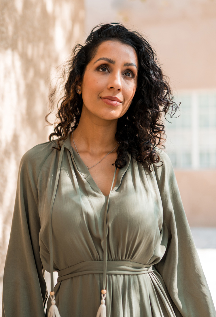 Portrait of a woman with curly hair wearing an olive-green dress, gazing thoughtfully outdoors in soft sunlight.