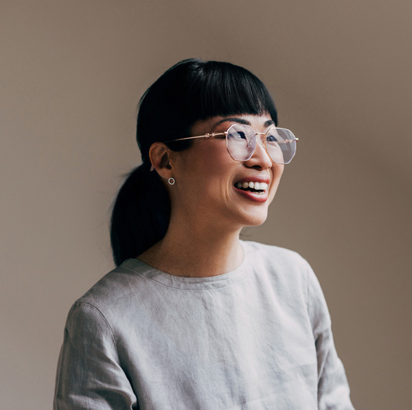 Portrait of a smiling woman with glasses and bangs wearing a gray blouse looking right on a neutral background