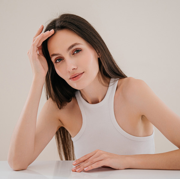 Young woman in a white tank top leaning on a table, relaxed expression and natural makeup