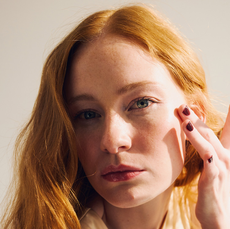 Close-up portrait of a red-haired woman with green eyes touching her cheek in warm natural light