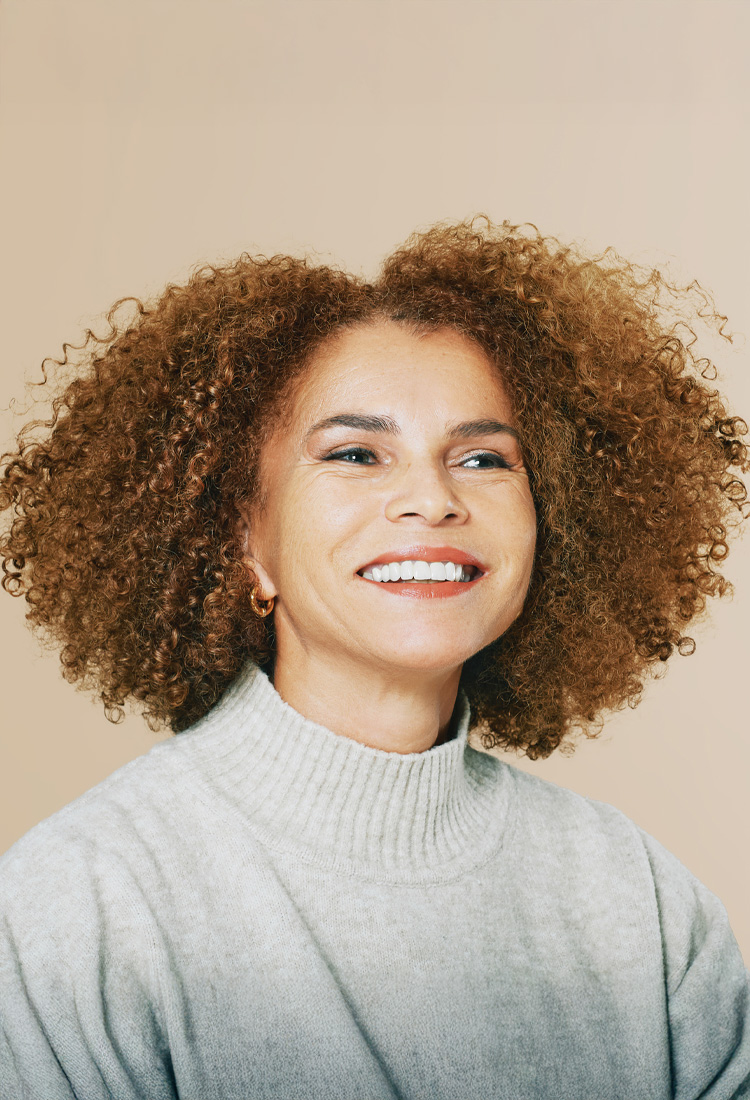 Smiling woman with voluminous curly hair wearing a light gray turtleneck sweater in a beige studio portrait