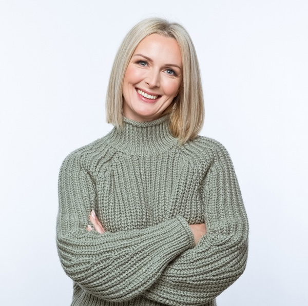 Smiling woman with blonde bob wearing chunky sage-green turtleneck sweater, arms crossed against white background.