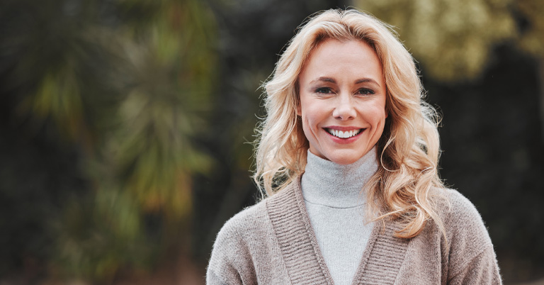 Smiling woman outdoors wearing a gray turtleneck and beige cardigan, casual portrait with blurred green background