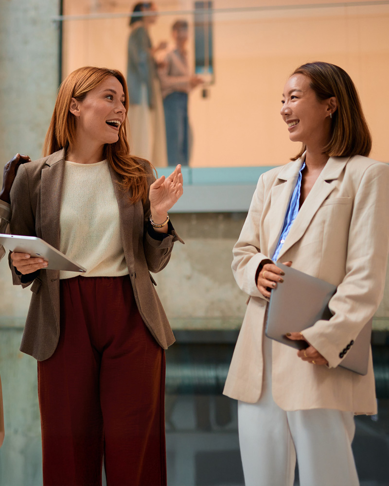 Two professional women smiling and talking in a modern office lobby, each holding a tablet or laptop.