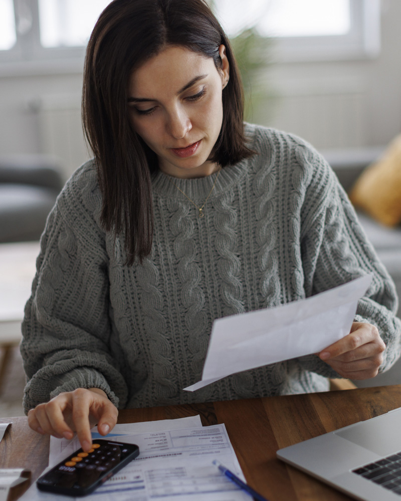 A woman calculating bills at home, reviewing paperwork with a calculator, laptop, and documents.