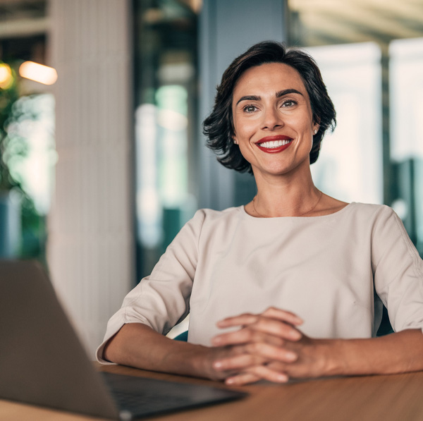 Smiling professional woman seated at desk with laptop in modern office, hands clasped, confident business portrait