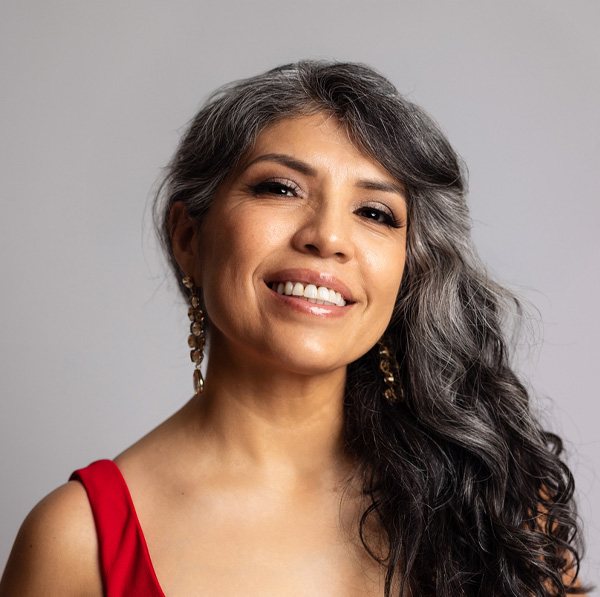 Smiling woman with long gray hair wearing a red dress and gold earrings against a neutral background