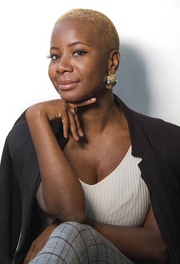 Portrait of a woman with short blond hair, gold earrings, white ribbed top and blazer, smiling with hand under chin.