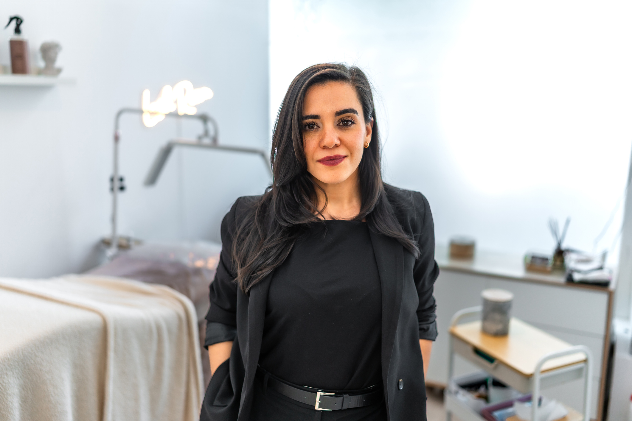 Confident med spa owner stands ready to treat any patient, with an exam table and light treatment tools behind her.